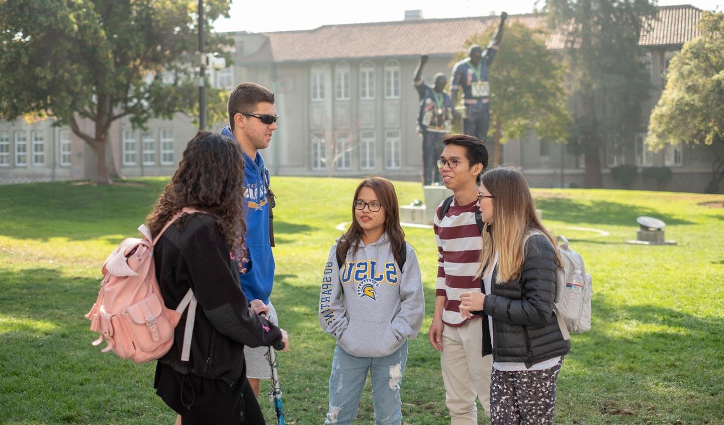 Five students talking on San José State University’s Tower Lawn with the Vicotry Salute monument in the distance.